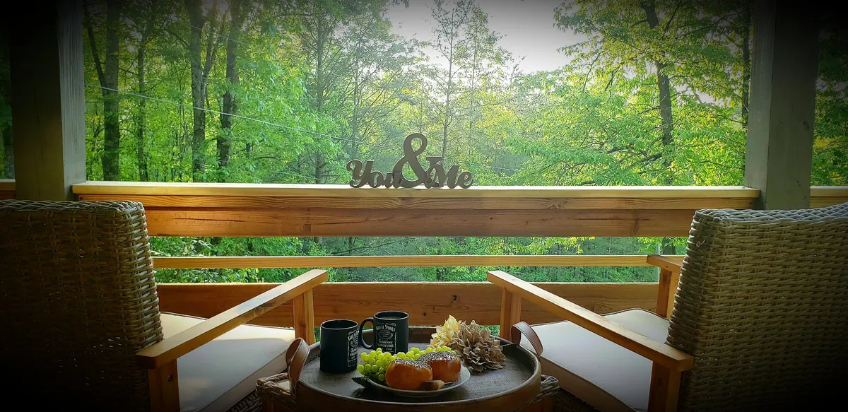 An inviting covered porch scene with comfortable chairs, a decorative 'You & Me' sign on the wall, coffee mugs, and a platter of fresh fruit and snacks, all set against a tranquil outdoor backdrop.