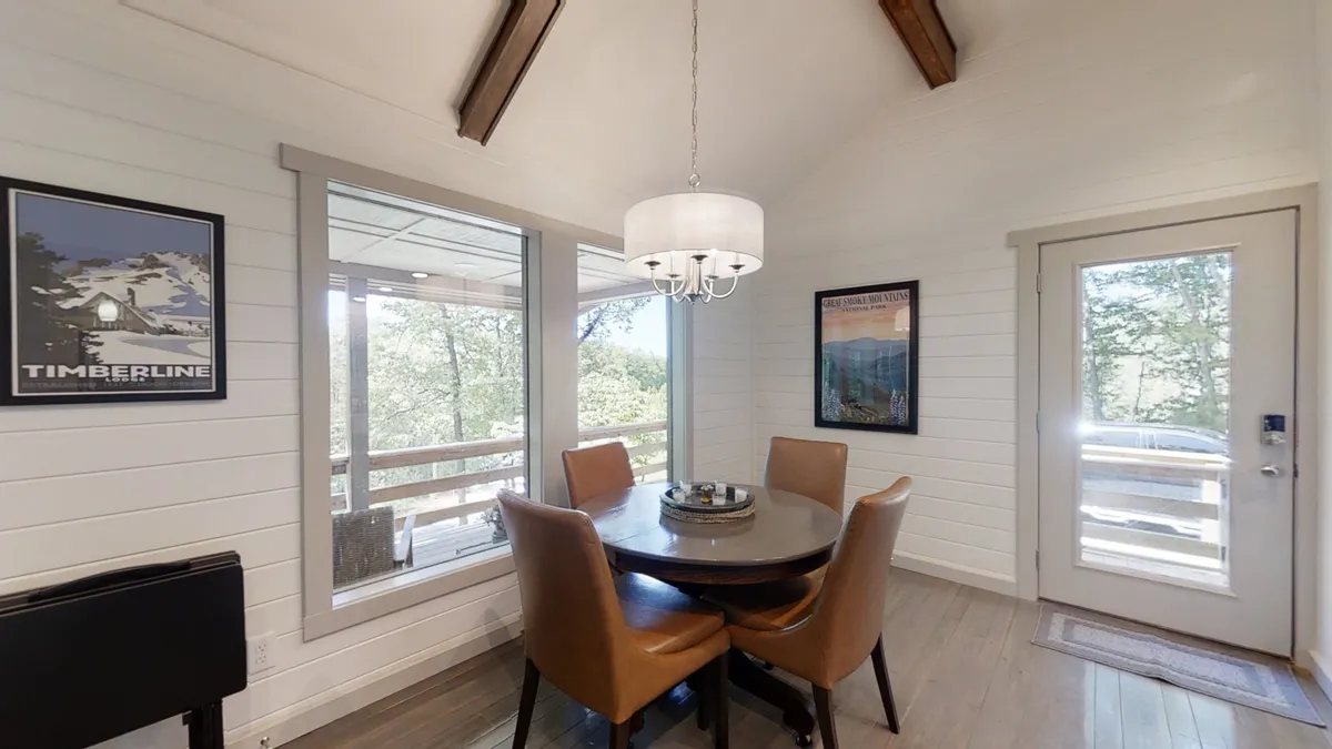 A cozy dining table sits beneath a chandelier, surrounded by windows and wall art. The arrangement evokes family meals, casual conversations, and holiday celebrations, all in a light-filled, welcoming space.
