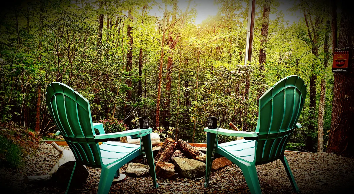 This image depicts two green Adirondack chairs arranged in front of a cozy fire pit, surrounded by tall trees. The scene is serene, bathed in the warm glow of firelight with hints of the sunset filtering through the branches, evoking a sense of tranquility and connection with nature.