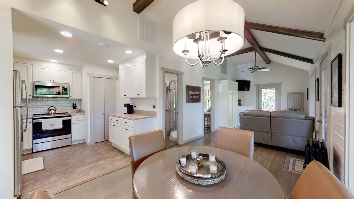 A cozy dining area set with a wooden table for four, surrounded by kitchen appliances and a welcoming living area. A chandelier above adds warmth and charm, making it ideal for family meals and holiday celebrations.