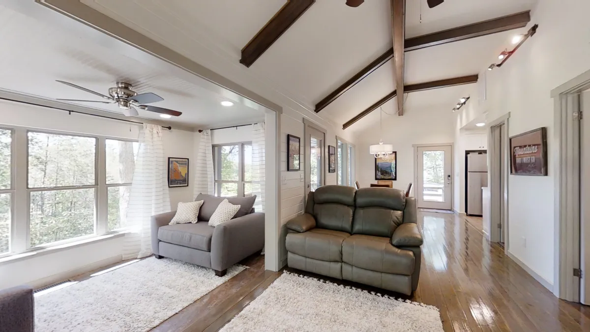 A comfortable seating area with expansive windows letting in abundant natural light and showcasing the wooded environment just outside. The wooden ceiling beams and cozy furniture create a welcoming retreat after a day of outdoor exploration.