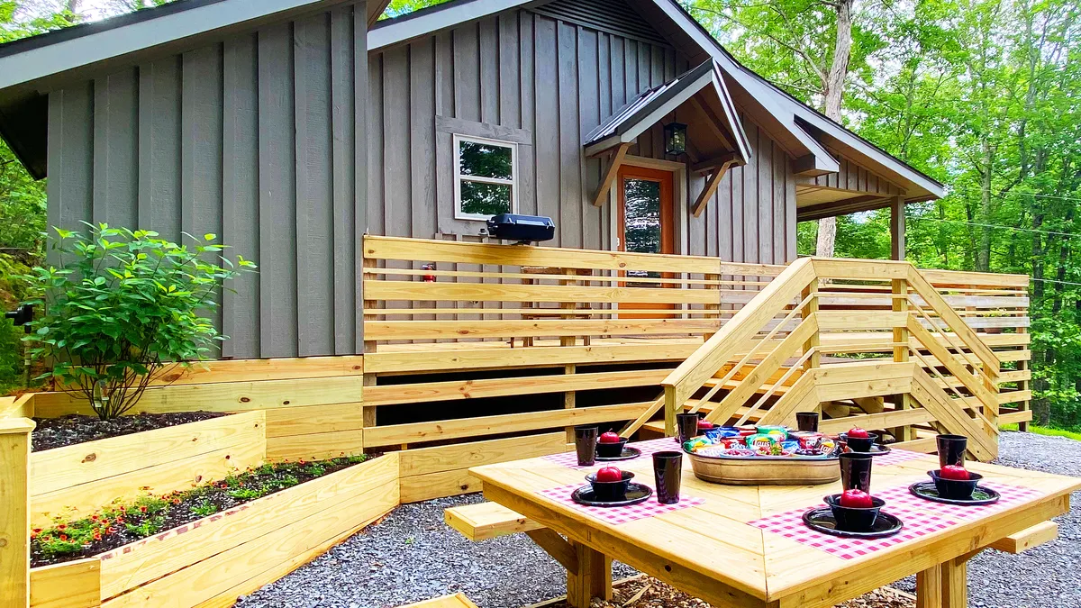 The image showcases the cottage's inviting wooden deck set for dining, surrounded by lush greenery. The modern cabin facade is visible in the background, with the deck positioned to take in views of nature, perfect for gatherings or peaceful relaxation. The scene is bathed in soft, golden light, suggesting sunset or early morning, setting a tranquil and welcoming mood.