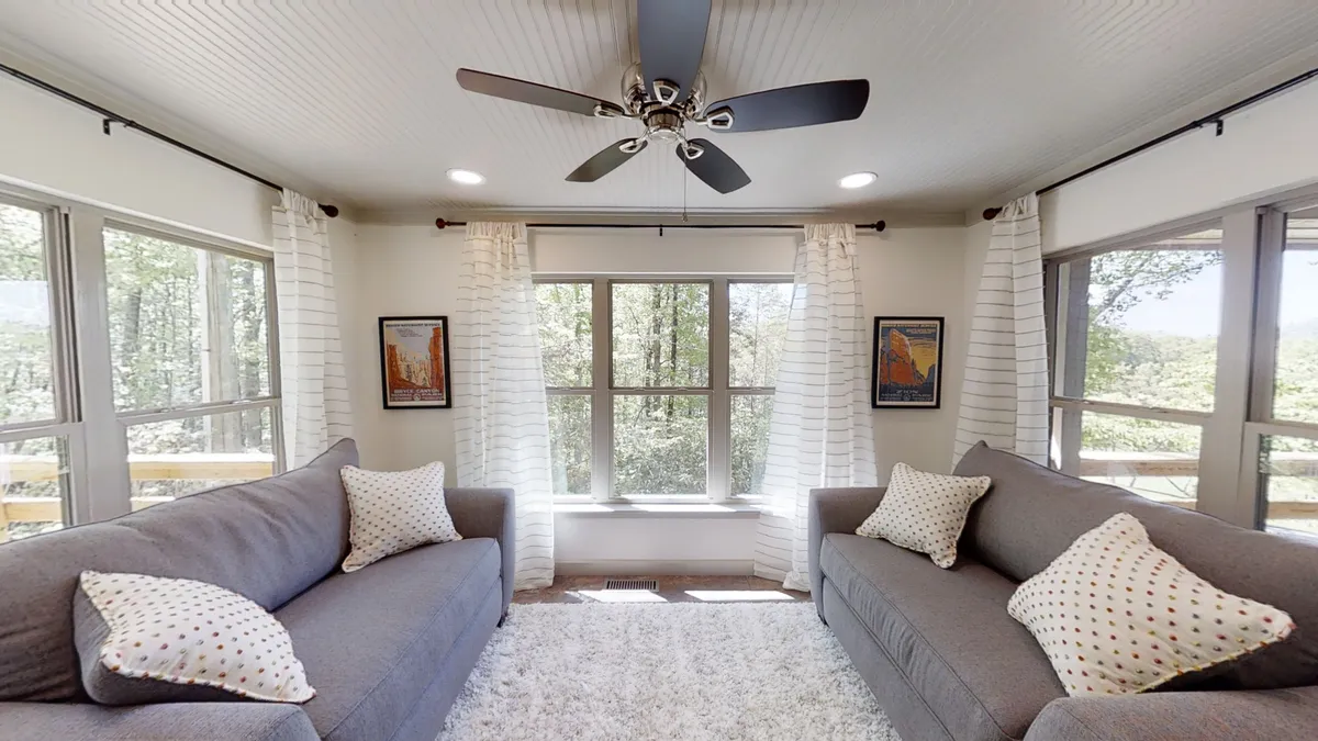 A spacious, sunlit seating area with plush sofas, large windows letting in ample natural light, a ceiling fan, and colorful decorative pillows. Artwork adorns the walls, and the room radiates comfort and invitation for relaxation or socializing.