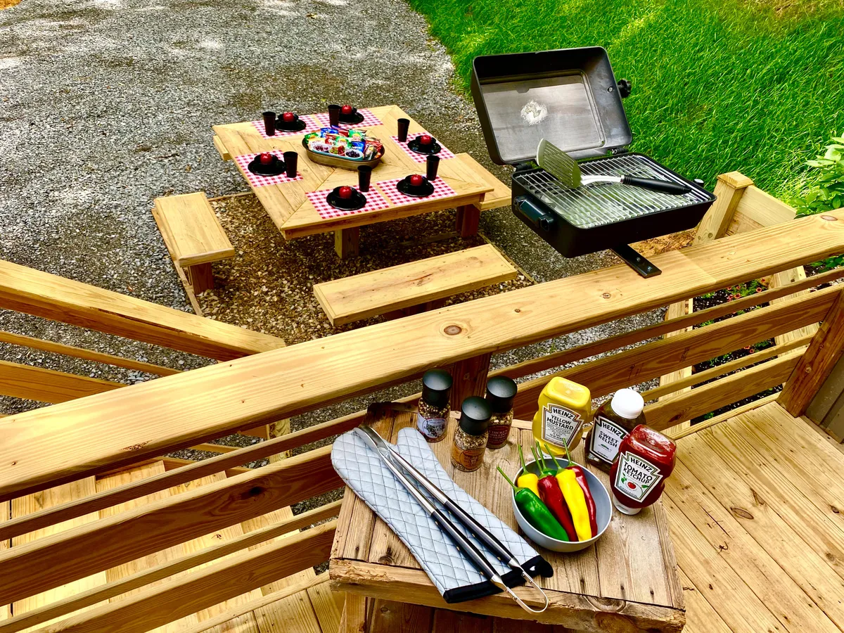 An outdoor picnic table and a gleaming gas grill set amid lush greenery, with condiments and fresh vegetables ready for a barbecue. The setup radiates a festive, casual vibe, ideal for al fresco meals under the trees.