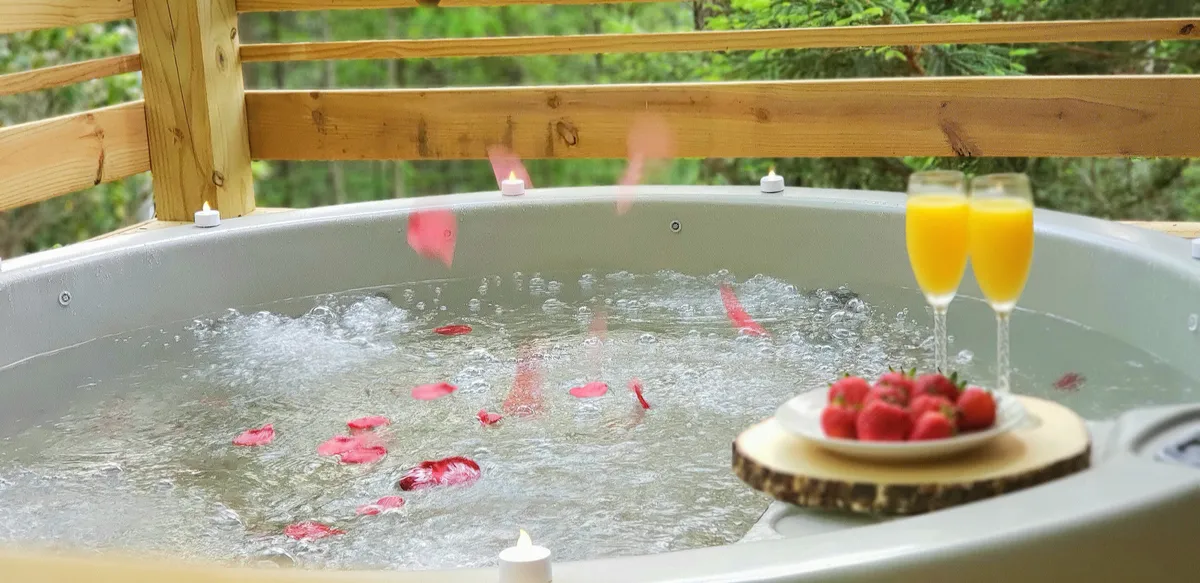 The image showcases a luxurious hot tub adorned with rose petals, flanked by glasses of champagne and fresh strawberries, all set against a backdrop of nature. This setting suggests relaxation and indulgence, with a special touch of celebration and intimacy.