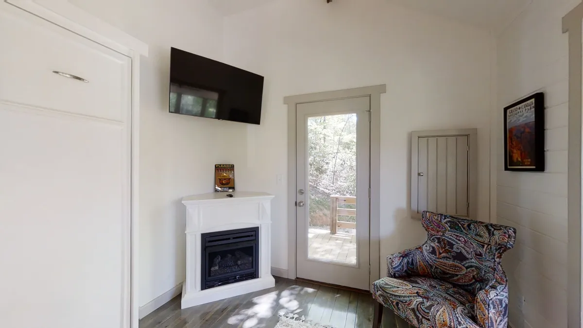 A warm and inviting living area with a fireplace, colorful accent chair, and a glass door leading directly to the outdoors. Through the door, you glimpse the greenery outside, hinting at the natural setting that surrounds the cottage.