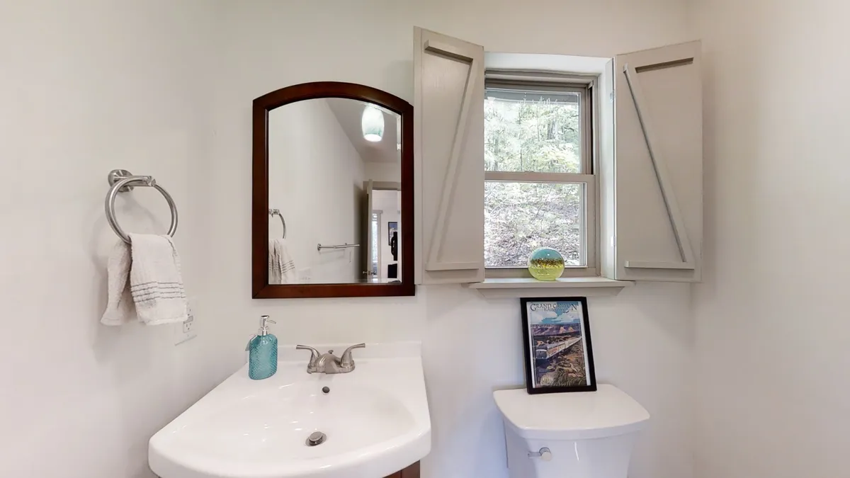A sunlit bathroom with a sink, mirror, and window with shutters. A decorative item sits on the sill, and a framed picture adds a personal touch. The window brings in natural light, offering a glimpse of the outdoors and a peaceful moment to start the day.
